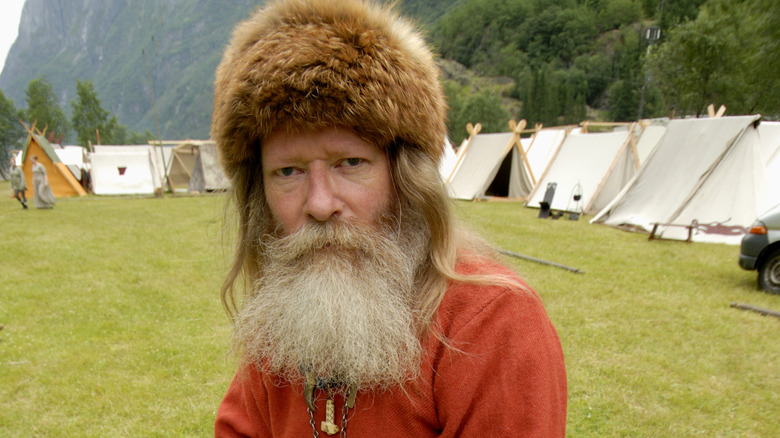 Un homme avec une longue barbe pâle et un chapeau de fourrure se tient devant des tentes viking traditionnelles.
