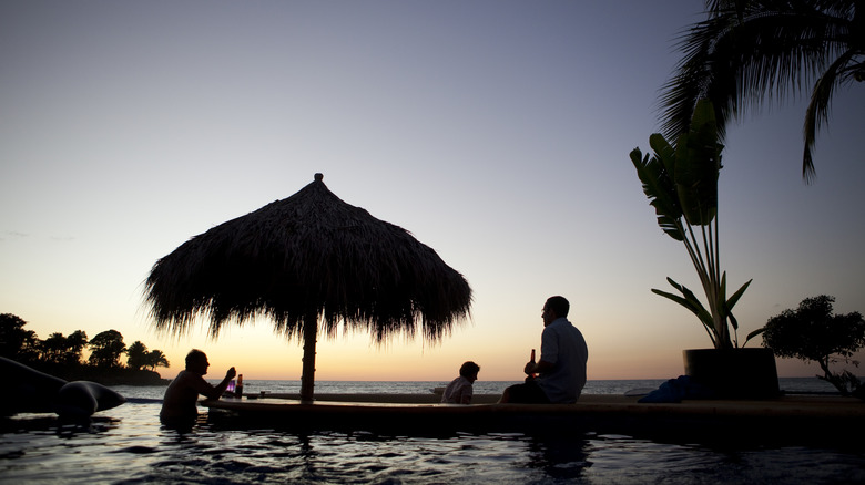Silhouette de personnes et un palapa par une piscine au coucher du soleil