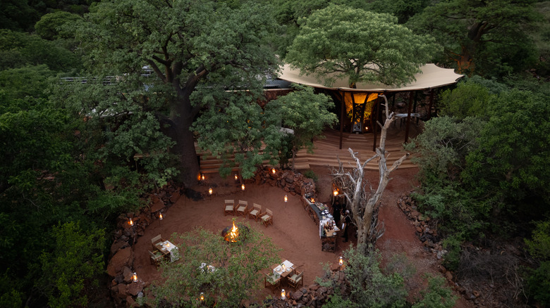 Des tables et des chaises entourant un feu de camp avec une table de nourriture installée à proximité, et une salle à manger principale derrière, toutes entourées d'arbres luxuriants et de rochers rouges.