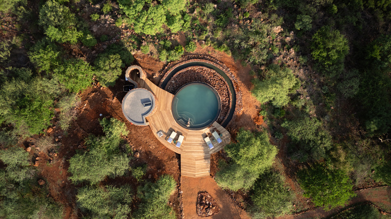 Vue aérienne d'une piscine isolée avec une plate-forme en bois entourée d'arbres et de rochers rouges avec une personne assise dans l'eau de la piscine.