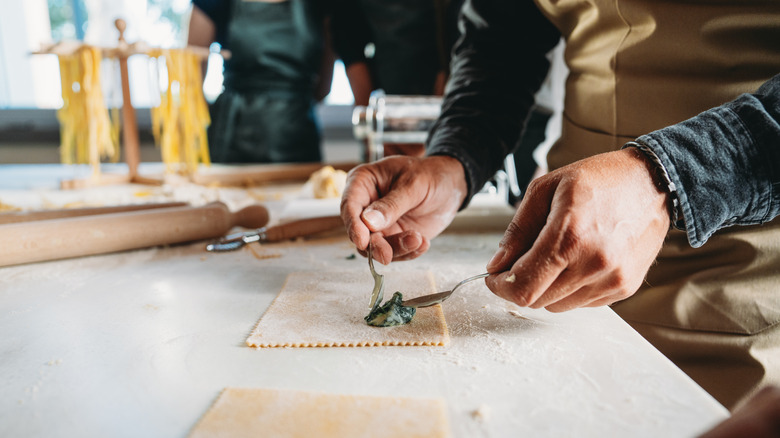 Les gens en cours de fabrication de pâtes entourant le comptoir avec des pâtes maison tandis que la personne place les épinards à la crème sur des raviolis