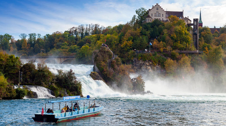 Un bateau touristique bleu s'approchant des chutes du Rhin en Suisse avec un château perché au-dessus sur une falaise