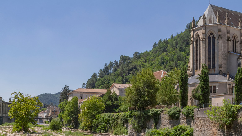 Église gothique près d'un lit de rivière rocheux, avec des maisons traditionnelles à proximité et entourées de verdure luxuriante, Vals-les-Bains