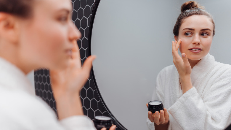Une jeune femme dans un peignoir se regarde dans le miroir tout en appliquant de la crème sur son visage