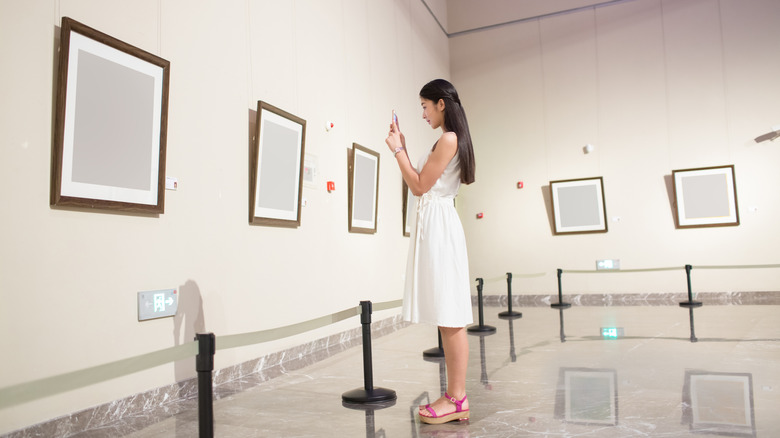 Une fille se tient devant des toiles vierges encadrées accrochées au mur d’une galerie et prend une photo avec son téléphone
