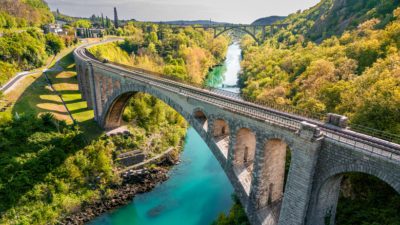 Un long pont de pierre avec des arches s'étend sur une rivière bleu-vert, avec une verdure luxuriante de chaque côté de l'eau.