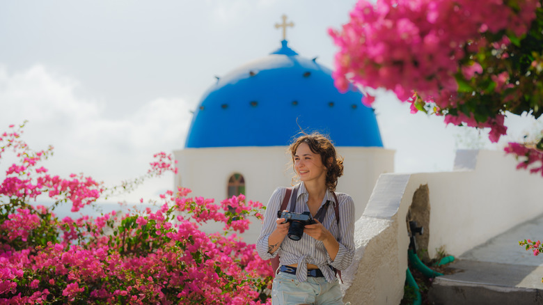 Une femme tenant un appareil photo avec une église au dôme bleu en arrière-plan, entourée de bougainvilliers en Grèce