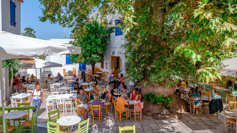 Une place principale animée avec des tables de restaurant sous un arbre contre des bâtiments blanchis à la chaux aux volets bleus