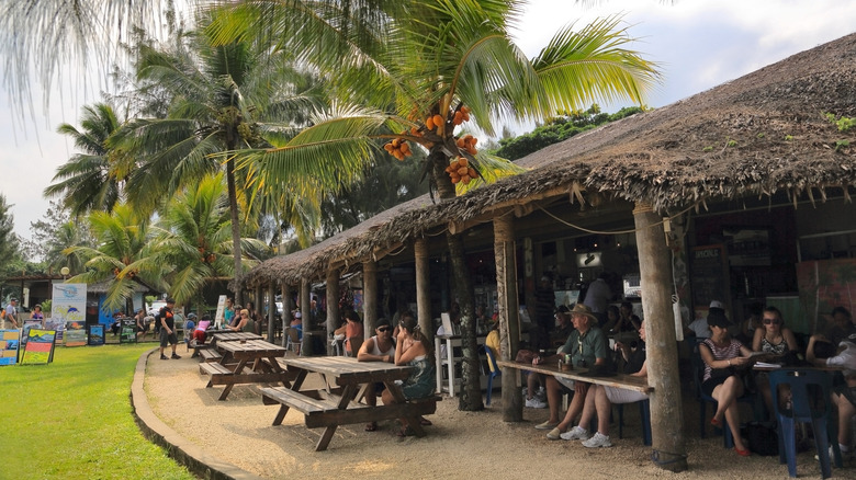 Touristes assis sur des bancs en bois sous un café au toit de chaume à l'ombre des palmiers
