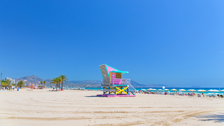 Ciel bleu sur une plage de sable blanc, avec un poste de secours coloré, des chaises longues et des palmiers