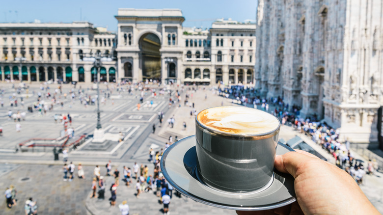 Une main tenant un cappuccino avec une place italienne animée en contrebas