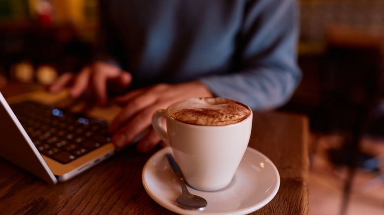Une paire de mains utilisant un ordinateur portable sur un bar avec un cappuccino