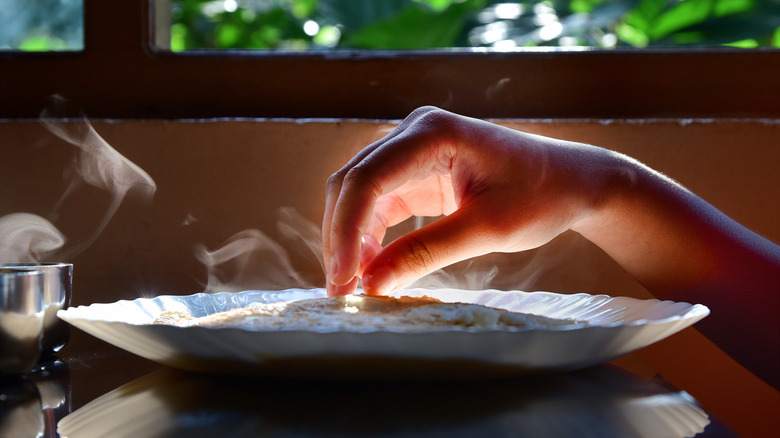 Gros plan de la main droite d'une femme cherchant un dosa, un pain plat indien généralement consommé au petit-déjeuner, servi sur une assiette en porcelaine, avec de la lumière traversant une fenêtre en arrière-plan